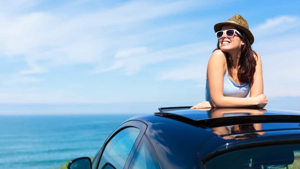 Relaxed happy woman on summer travel vacation to the coast  leaning out car sunroof with the sea on background.